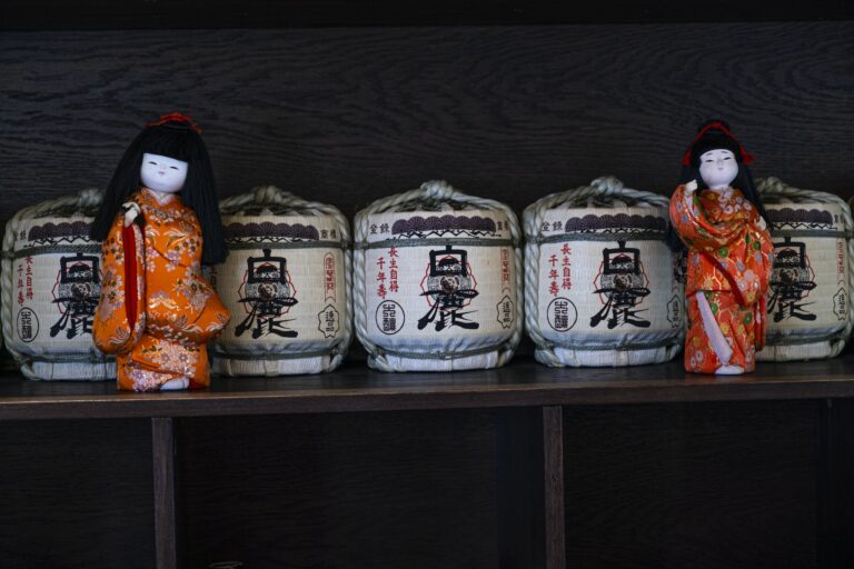Traditional Japanese Kimekomi dolls in red kimonos placed on shelf with abundance of wooden sake barrels with ropes and hieroglyphs